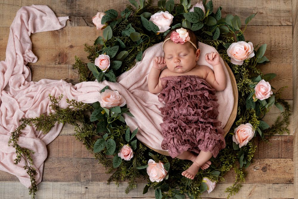 Newborn sleeping peacefully in plum ruffled dress surrounded by pink roses and greenery on rustic wooden floor.