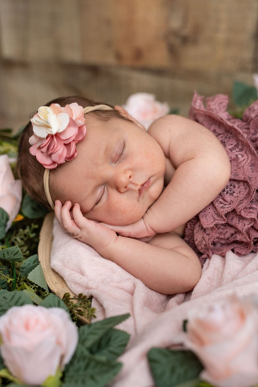 A sleeping newborn wearing a pink floral headband lies peacefully surrounded by pink roses and greenery.