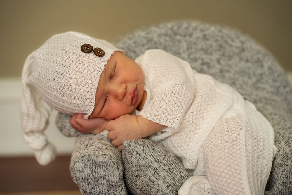A peacefully sleeping newborn wearing a cozy white knit outfit rests on a gray textured surface.