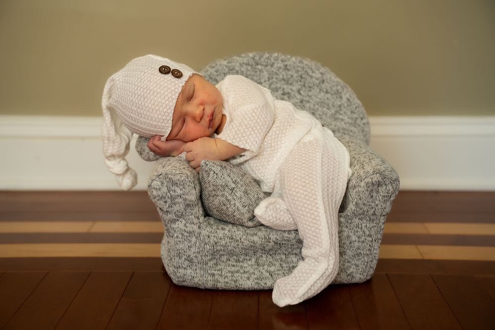 A newborn baby wrapped in white knit swaddle sleeping peacefully on a plush gray armchair.