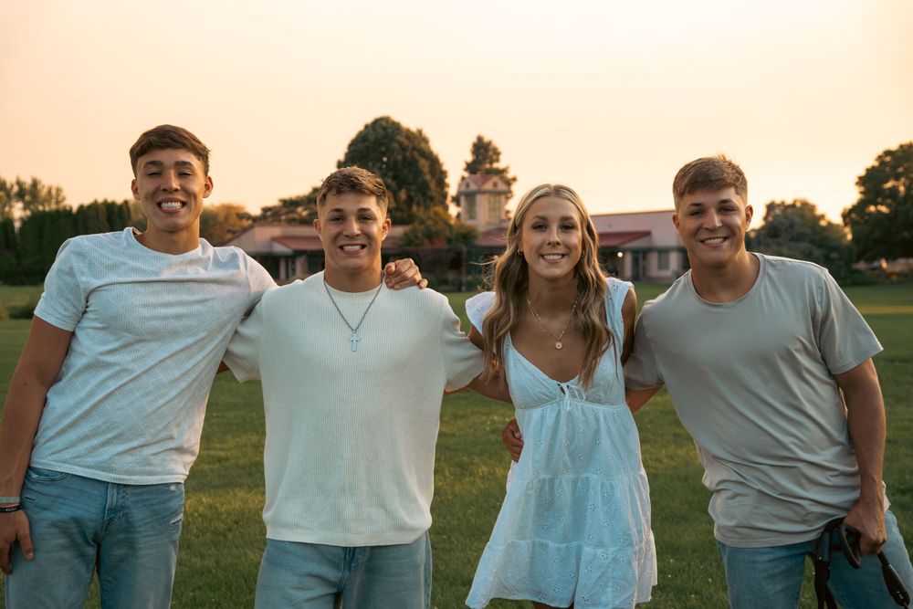 Four smiling teenagers posing together on green field at sunset in casual summer outfits.