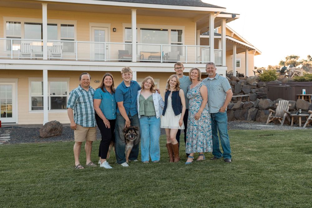 A joyful family of eight poses together on a green lawn in front of a yellow two-story home, dressed in coordinating teal outfits.