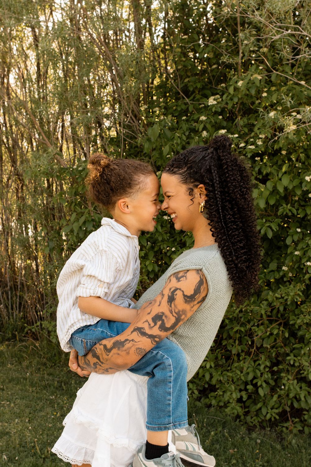A tattooed mother holds her young daughter, both smiling face-to-face outdoors among green trees.