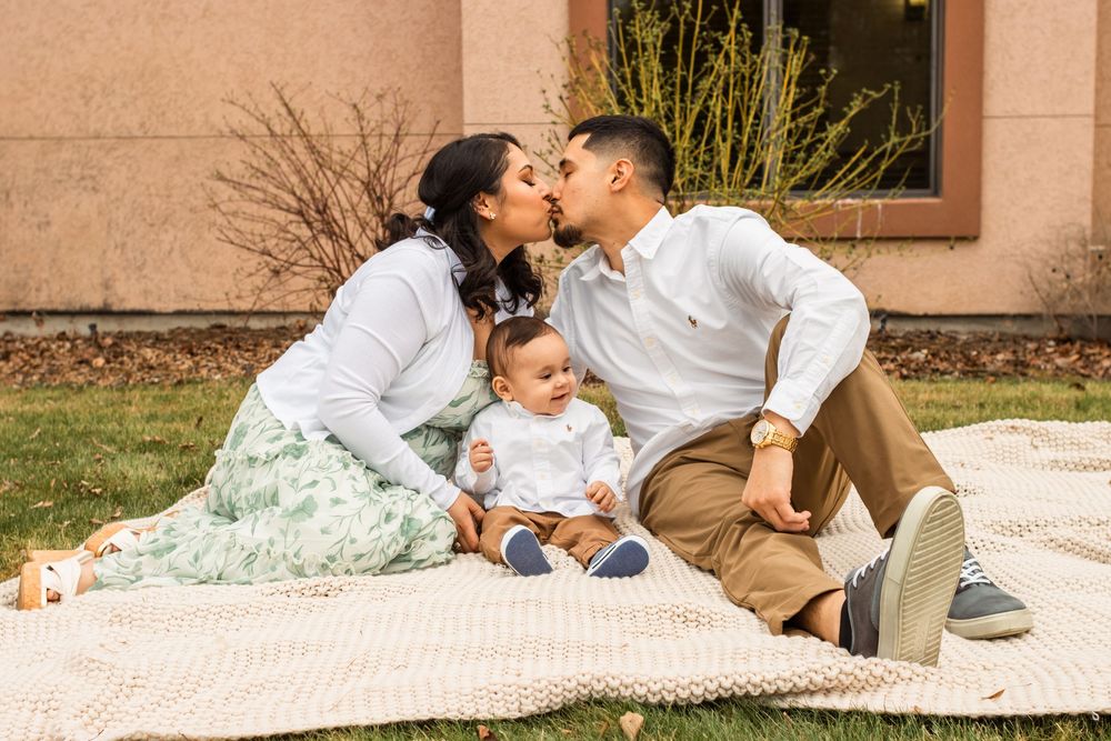 Young family sitting on blanket outdoors, parents kissing over smiling baby in white outfit.