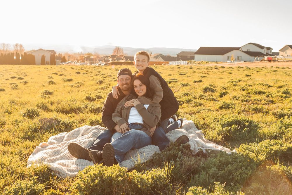Family of three sitting on a blanket in a sunny field, smiling warmly in golden hour light.