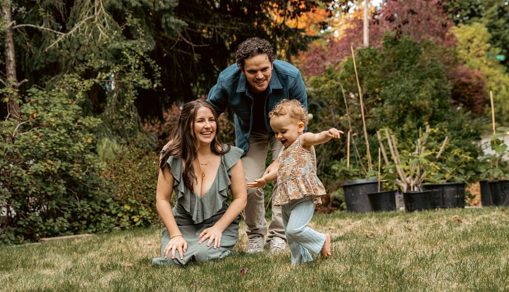 A happy young family plays together in a lush green garden with colorful autumn trees.