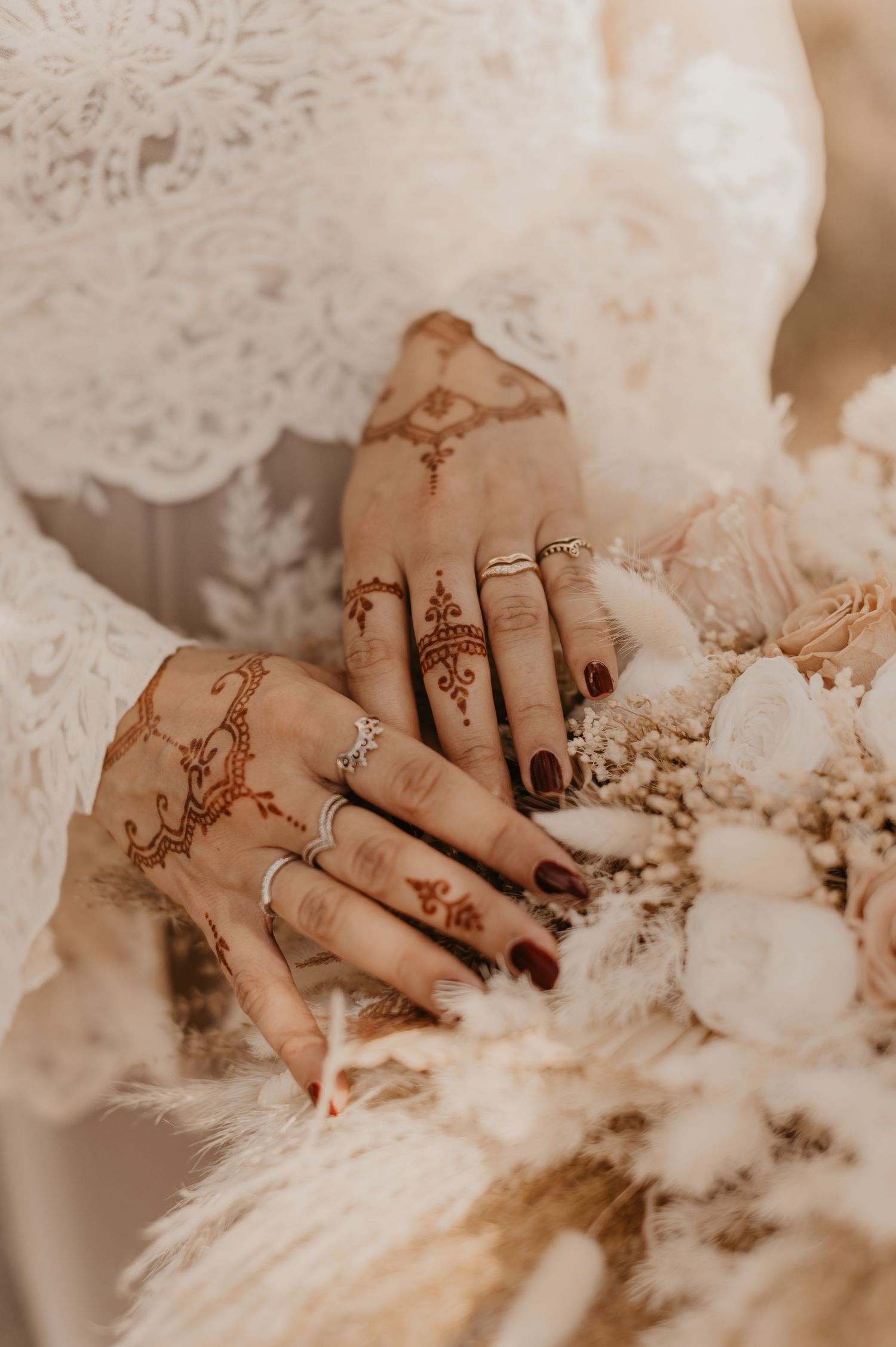 Close-up of hands adorned with intricate henna designs and dark nail polish against white lace and floral background.