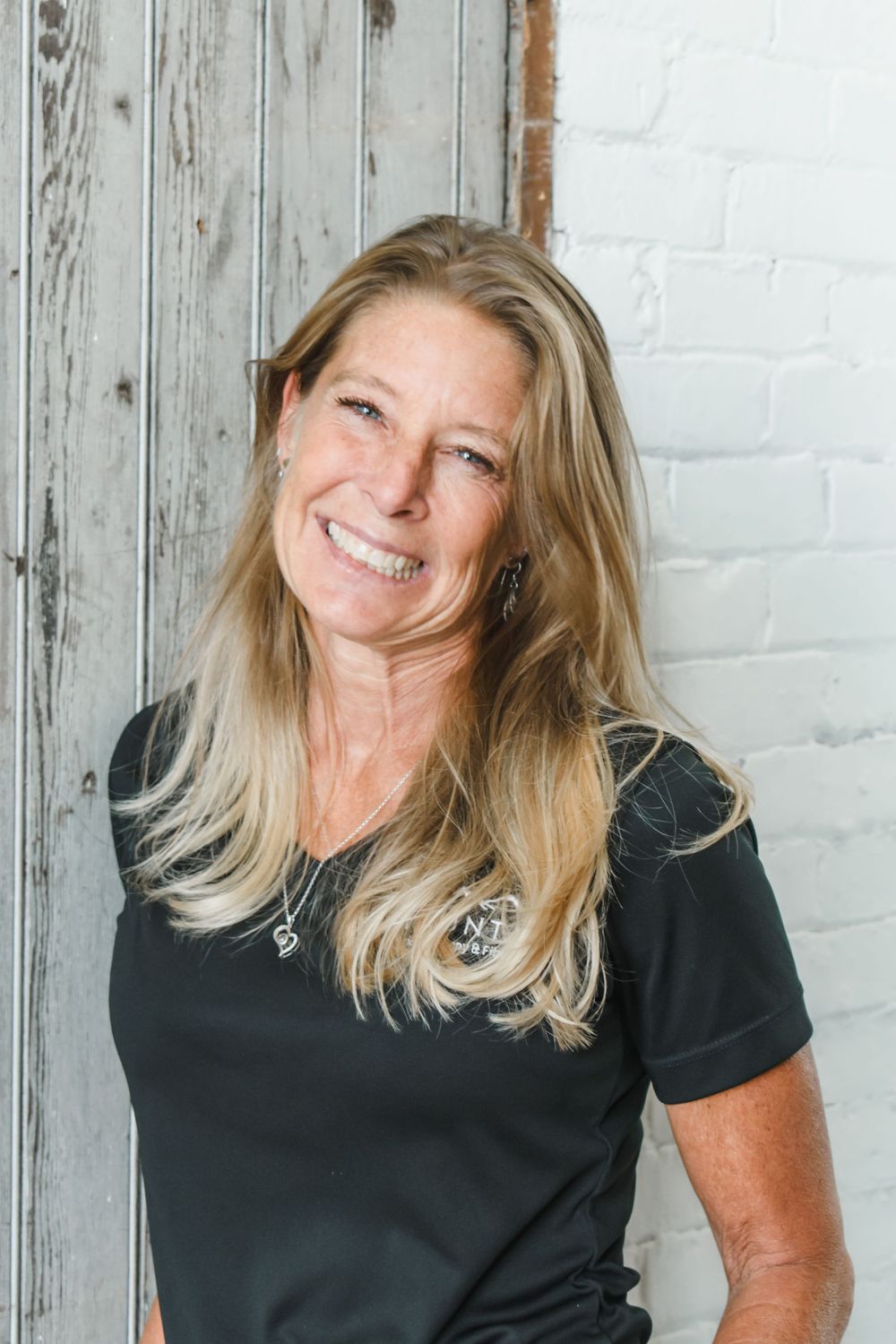 A joyful woman with long blonde hair wearing a black t-shirt poses against a rustic white wall.