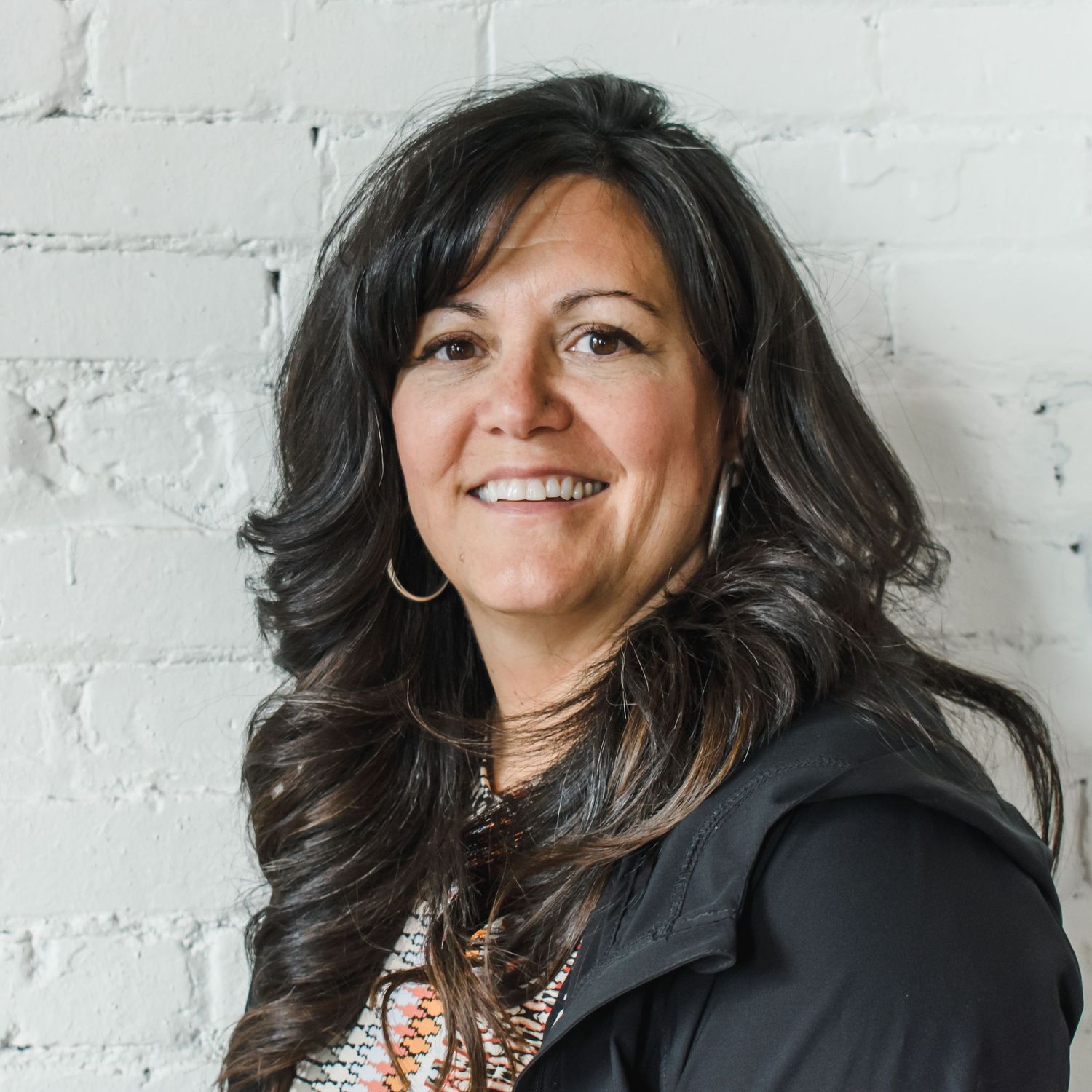 Professional headshot against white brick wall showing warm smile and long dark wavy hair.
