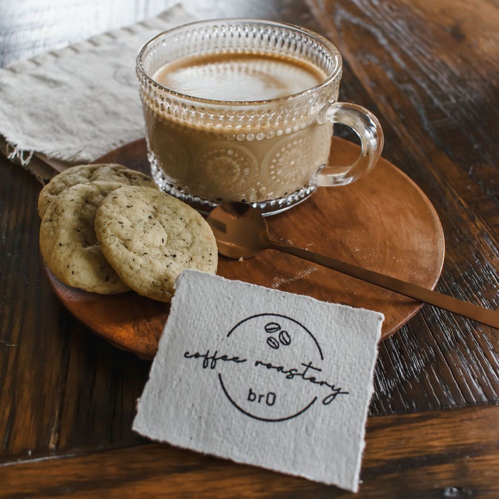 A glass mug of coffee with chocolate chip cookies on a wooden board next to a small paper card with coffee shop branding.