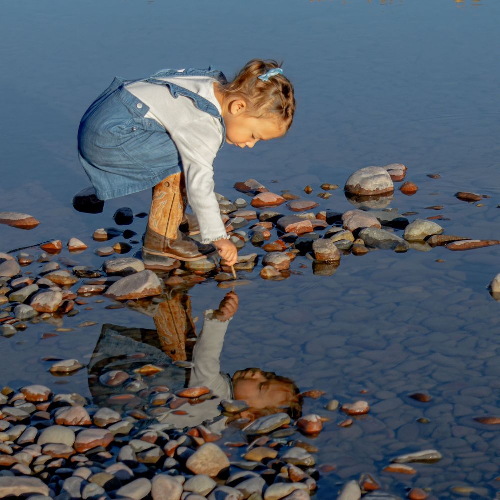 Person in white shirt bends down to examine rocks along a tranquil shoreline during sunset.
