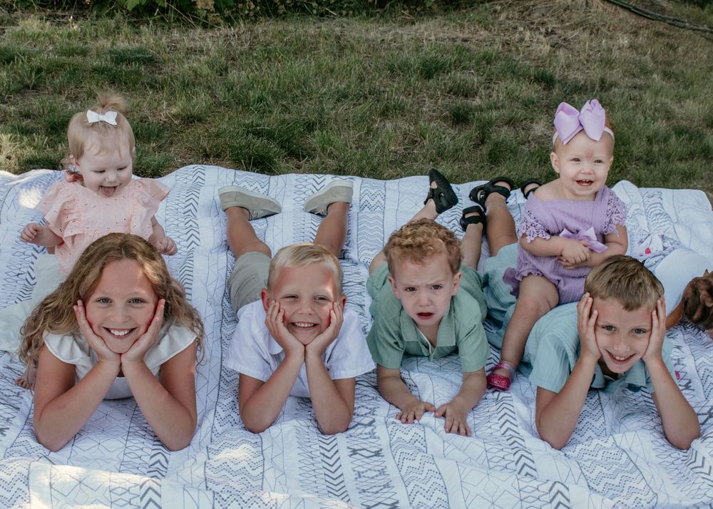 A series of children lying on a patterned blanket outdoors, posing with hands framing their faces in a playful manner.