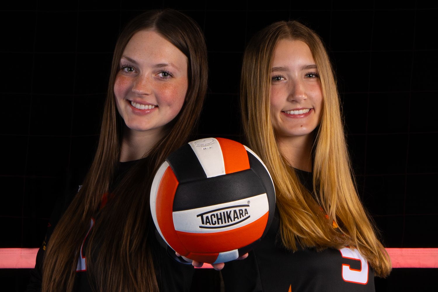 Two volleyball teammates pose with Tachikara volleyball against black backdrop.