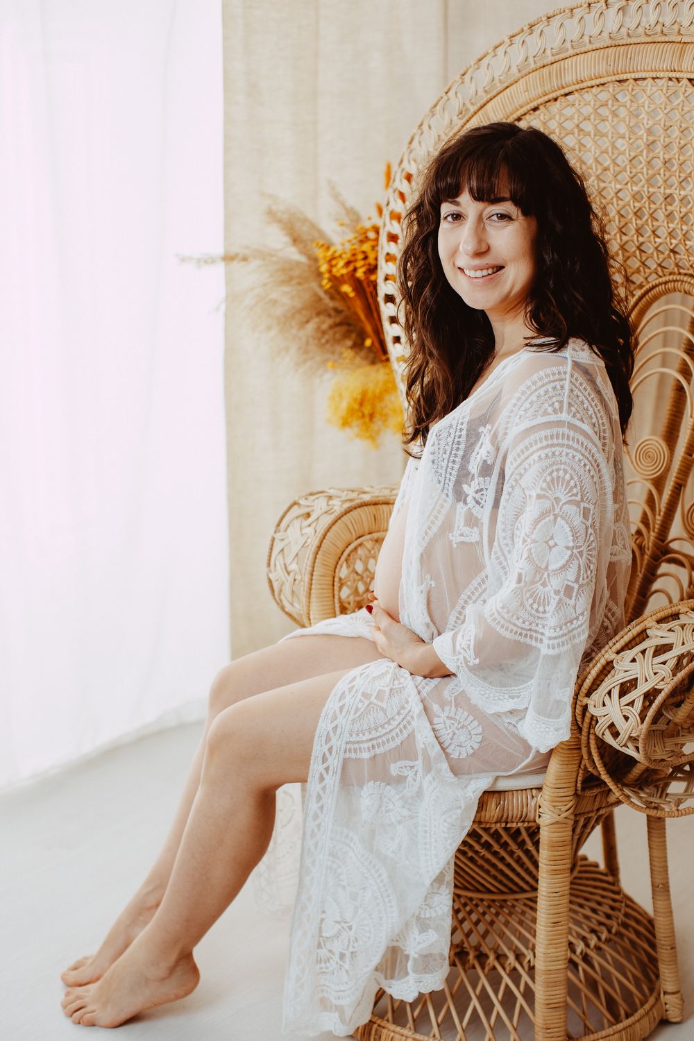 A person in a white lace dress sits barefoot in a vintage peacock chair with dried flowers in the background.