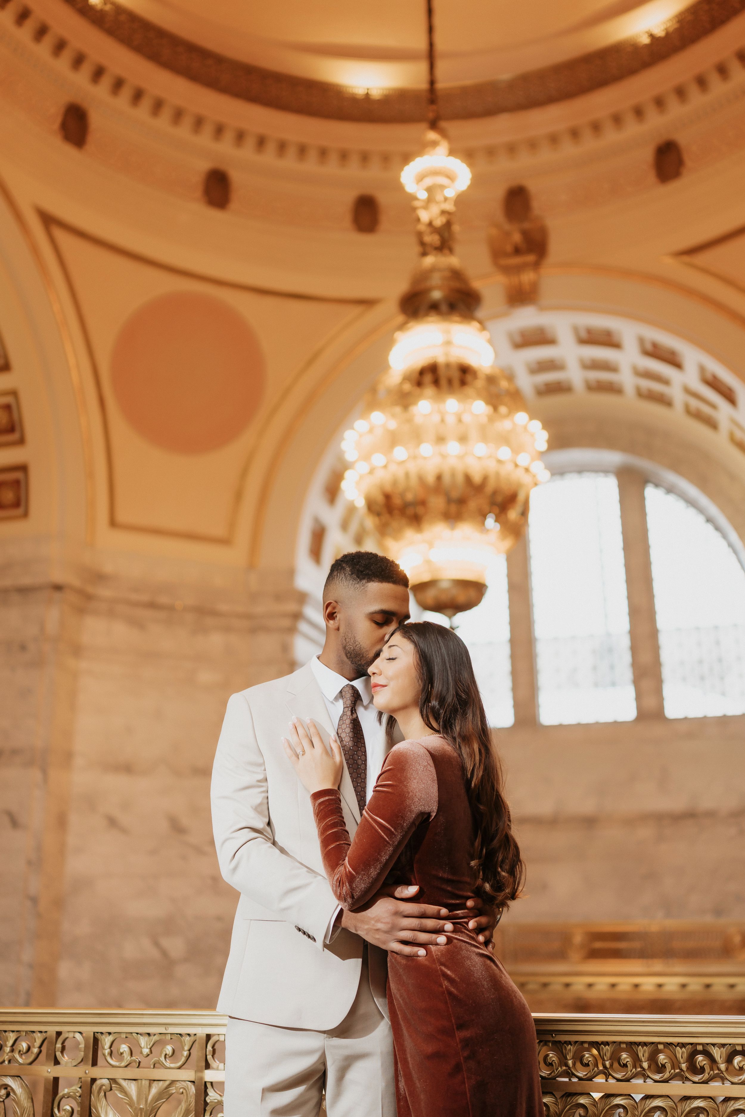 Romantic couple embraces under ornate chandelier in elegant historic building with architectural details.
