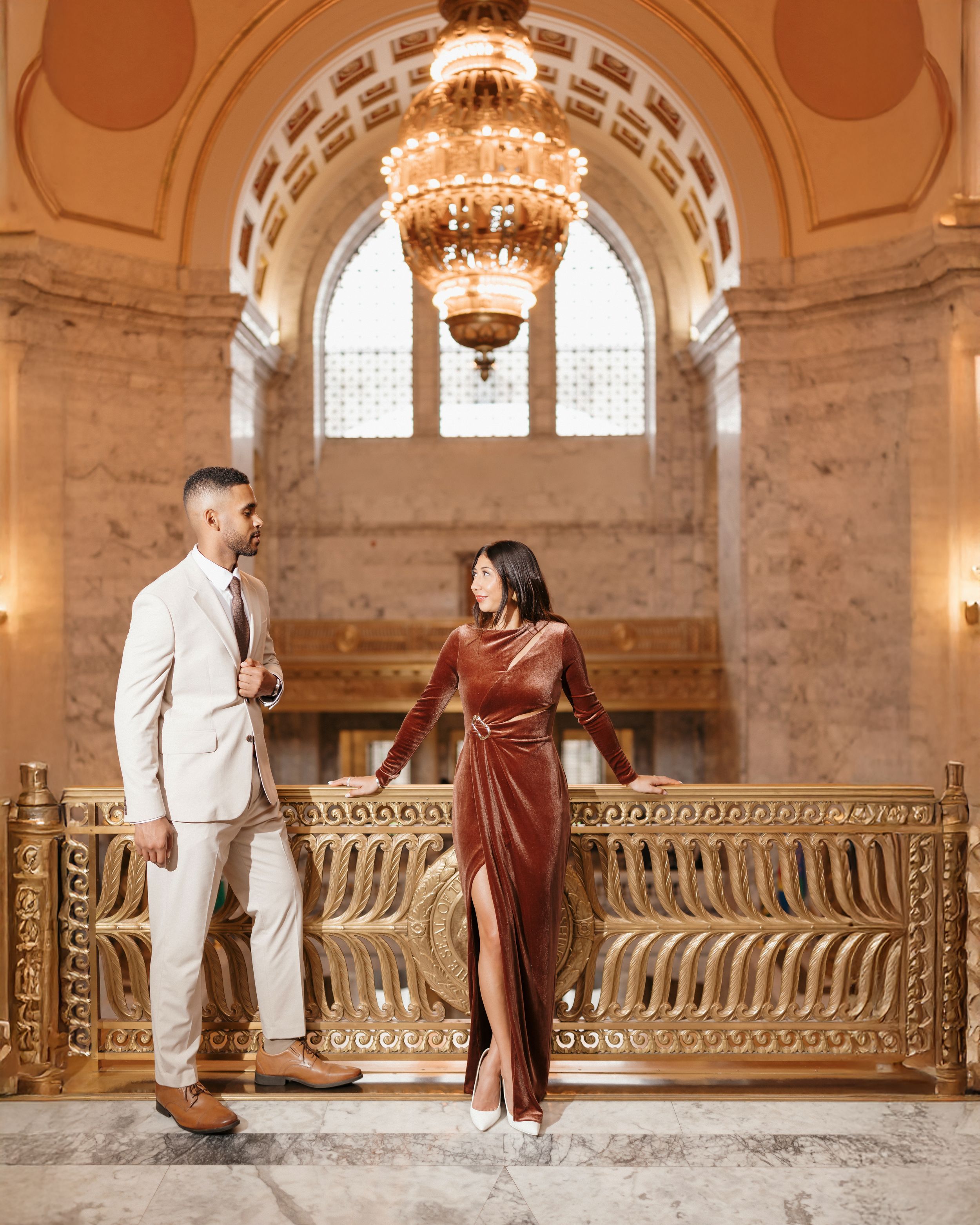 A couple in white and brown outfits poses together on a grand marble staircase with ornate railings in an elegant building.