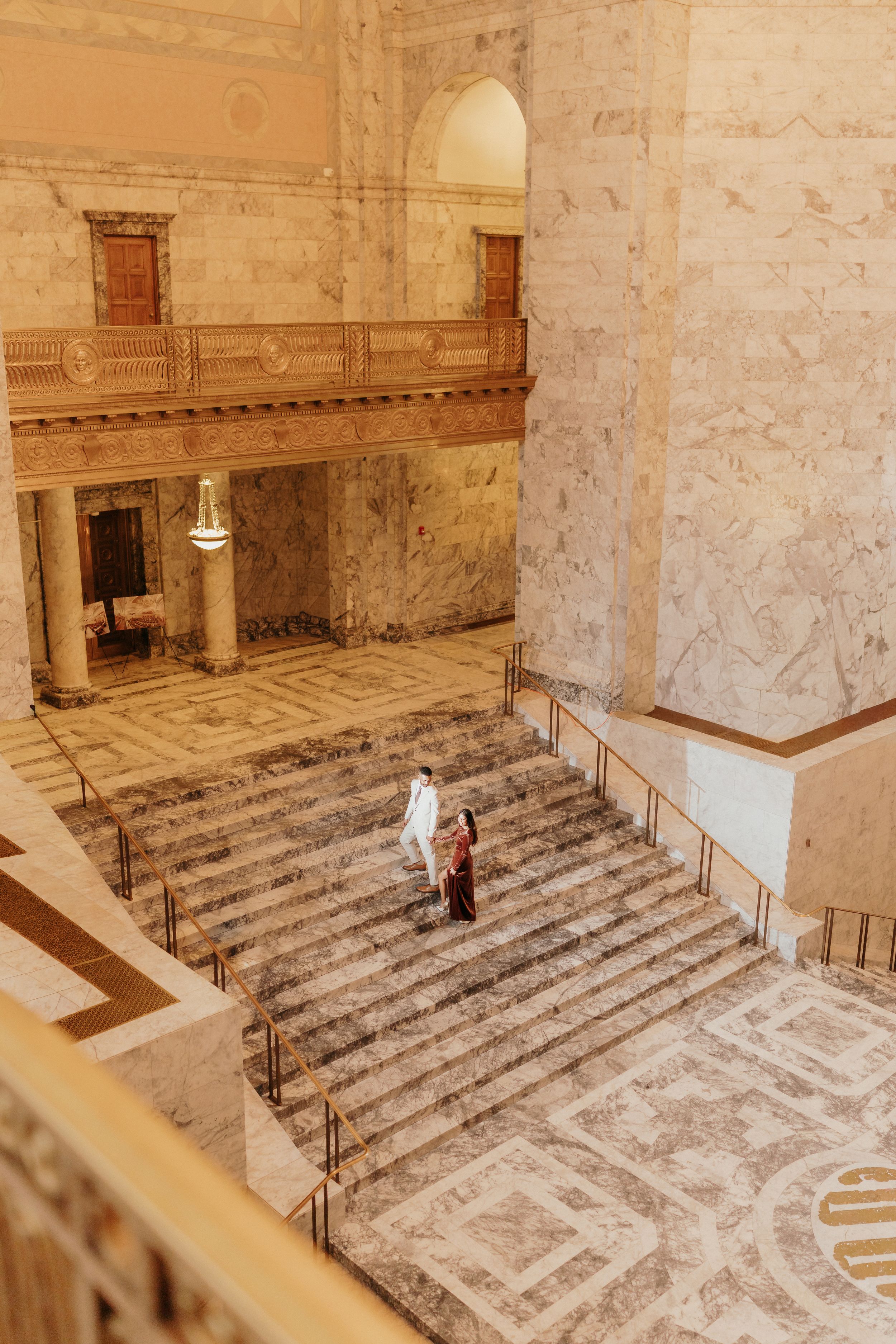A person in white stands on ornate marble steps inside a grand historic building with classical architectural details.