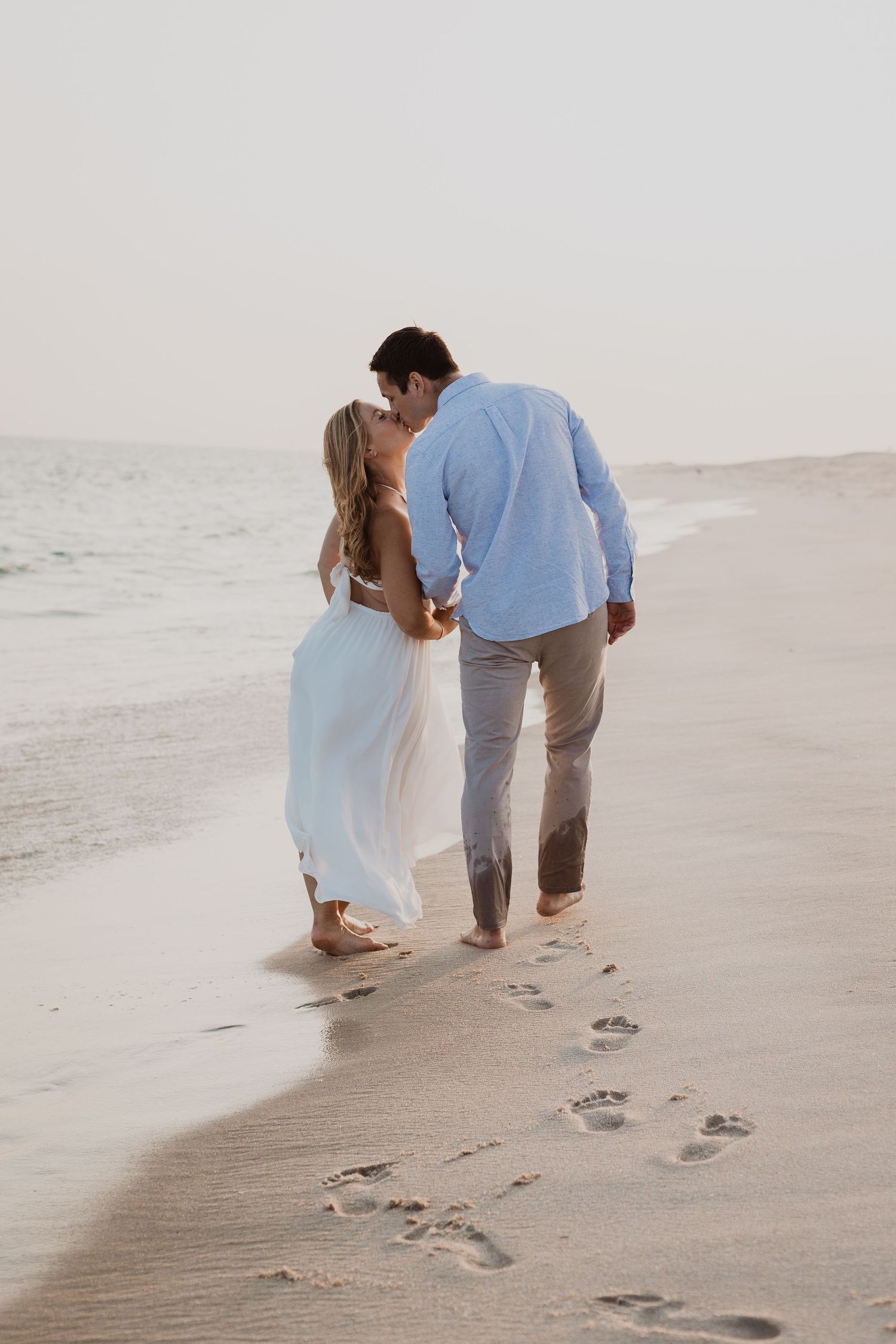 A couple in light blue and beige clothing walks along a sandy beach leaving footprints by the ocean shore.