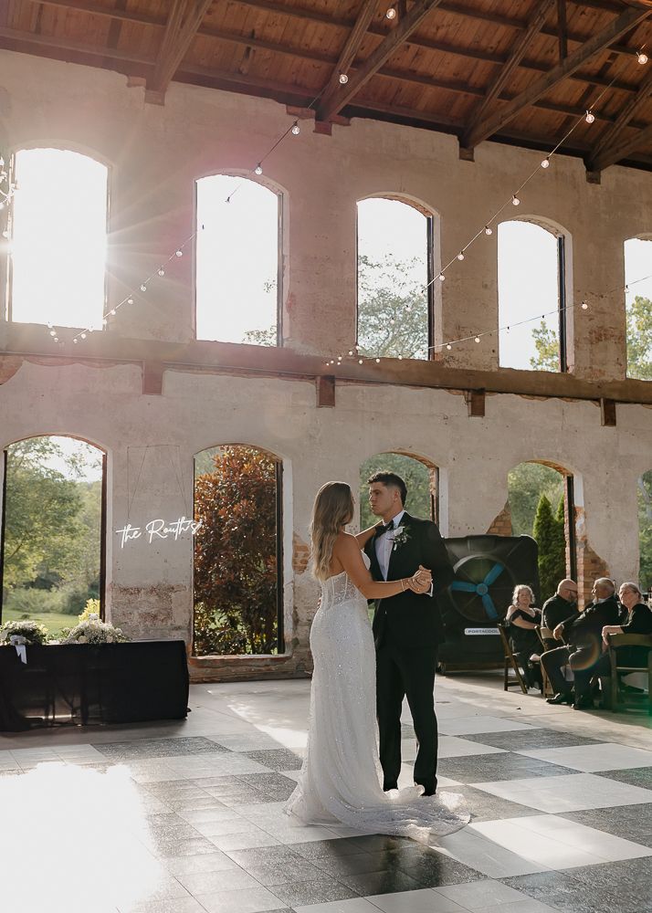 Wedding couple dances in a rustic venue with exposed wooden beams and arched windows.