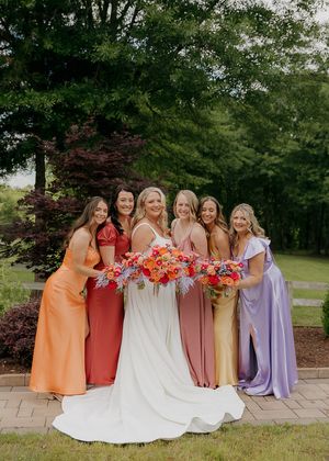 Bridesmaids in pastel orange, pink, and purple dresses pose together outdoors.
