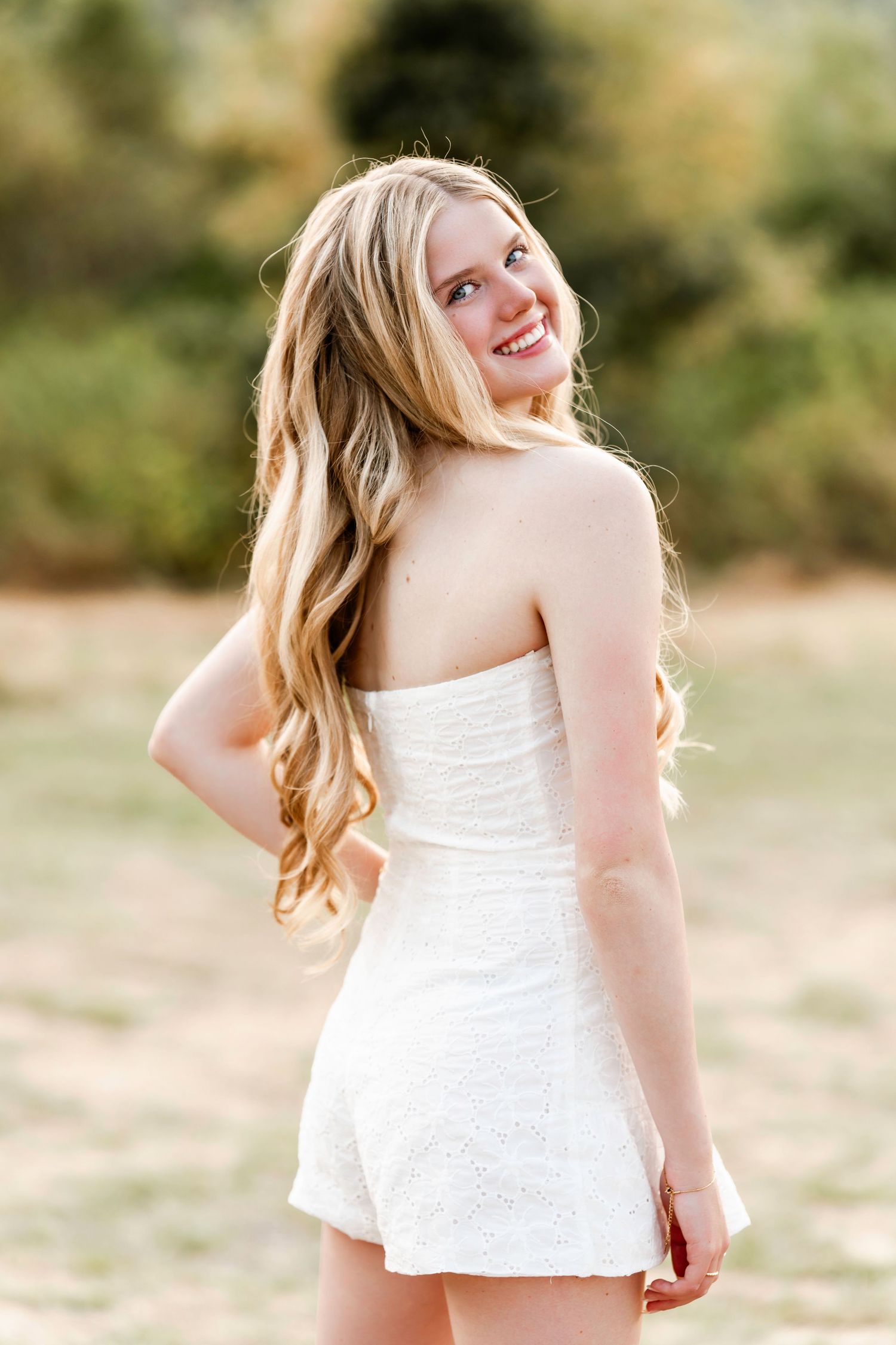High school senior smiling in a white lace romper during a golden hour outdoor session – natural and timeless senior photography.