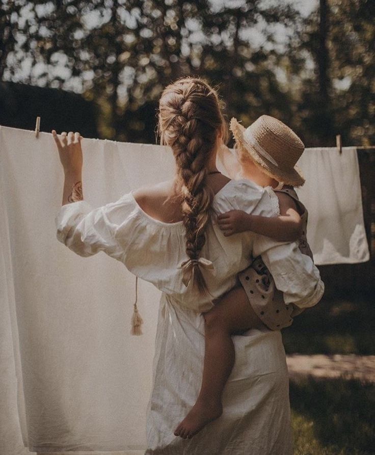 A woman with a long braid in a white off-shoulder dress holds a child near a clothesline outdoors.