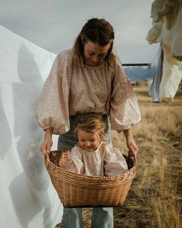 Mother and toddler in matching floral blouses, child sitting in wicker laundry basket outdoors near hanging white sheets.