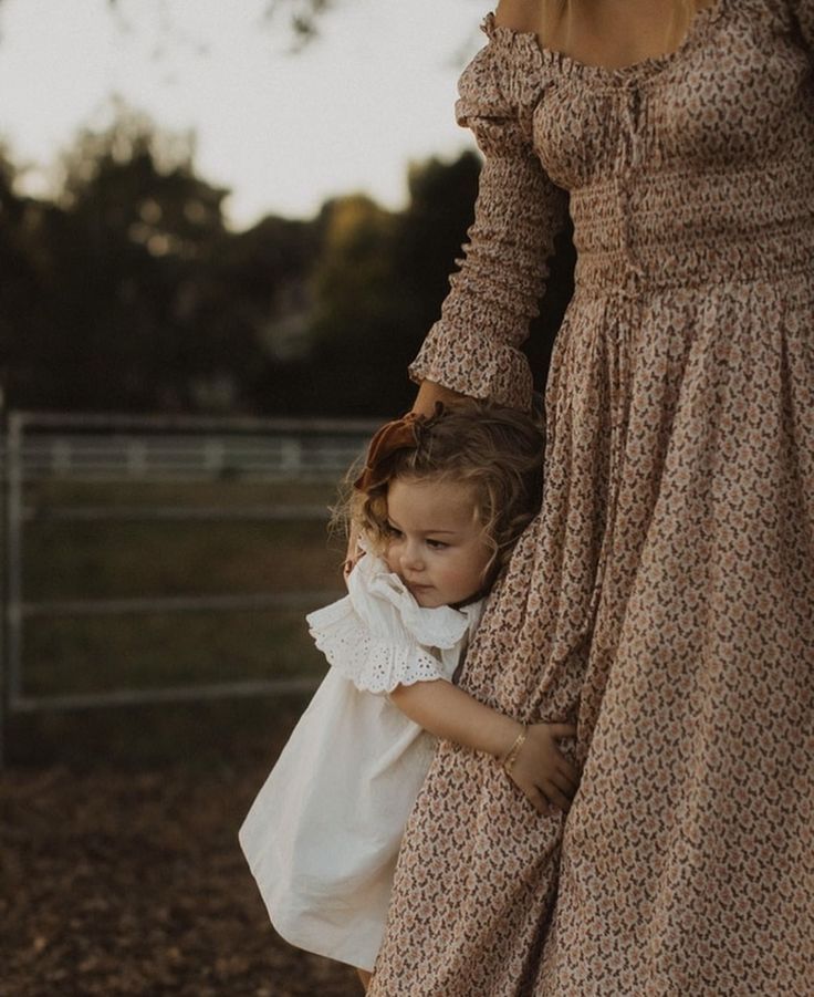 A toddler in a white dress hugs her mother's floral maxi dress outdoors at golden hour.