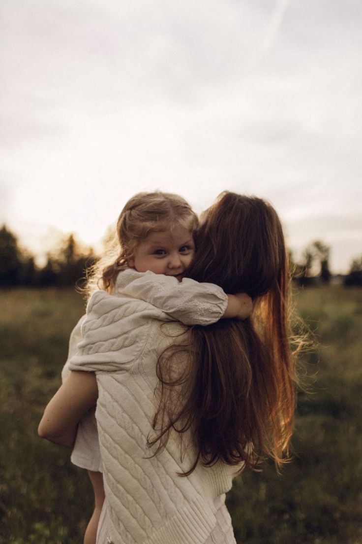 A young girl hugs a woman with long hair in a field at sunset, both wearing white knit sweaters.