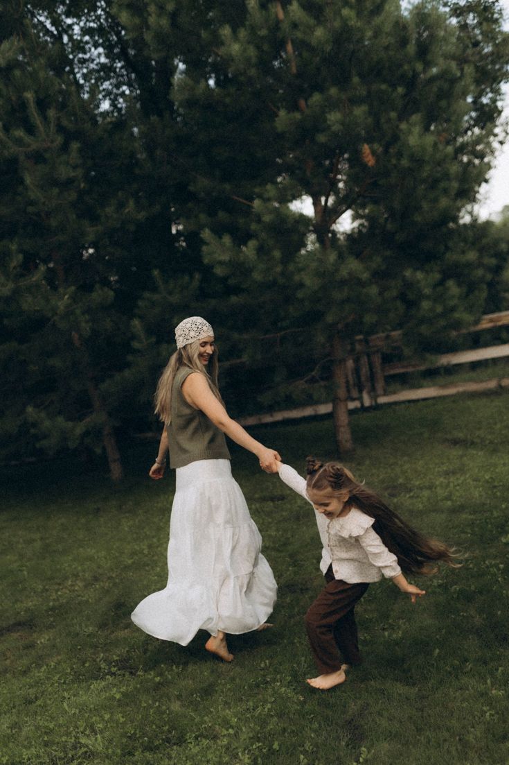 A mother and young daughter joyfully hold hands and spin together on green grass with pine trees in the background.