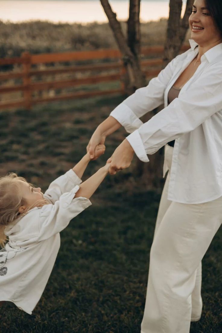 A joyful mother in white outfit spins her laughing blonde child outdoors near a wooden fence at dusk.