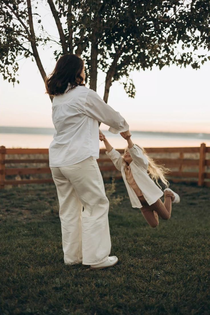 A mother in white outfit joyfully swings her blonde daughter outdoors near a wooden fence at sunset.