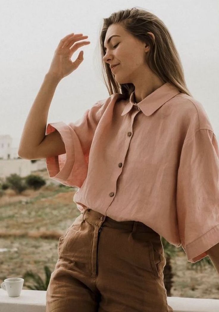 Woman in oversized pink linen cropped shirt and brown high-waist pants posing outdoors in natural light.