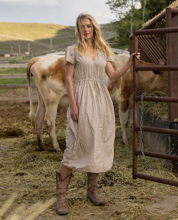 Woman in a floral midi dress and cowboy boots standing near cattle on a farm, rustic country style outfit.