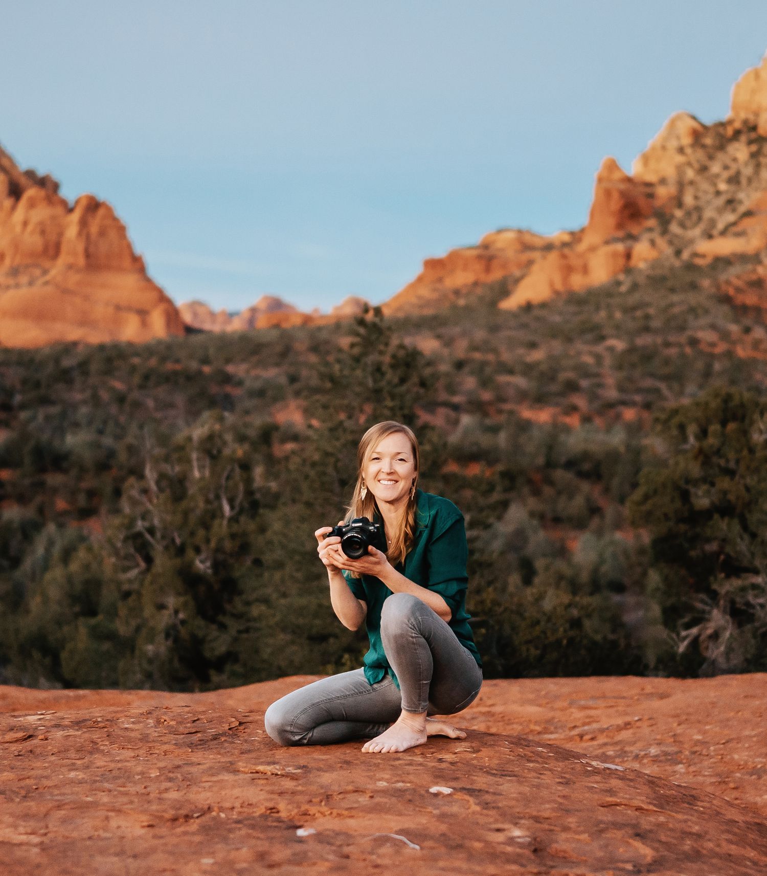 Smiling blonde photographer crouching on red rock with camera in Sedona landscape.