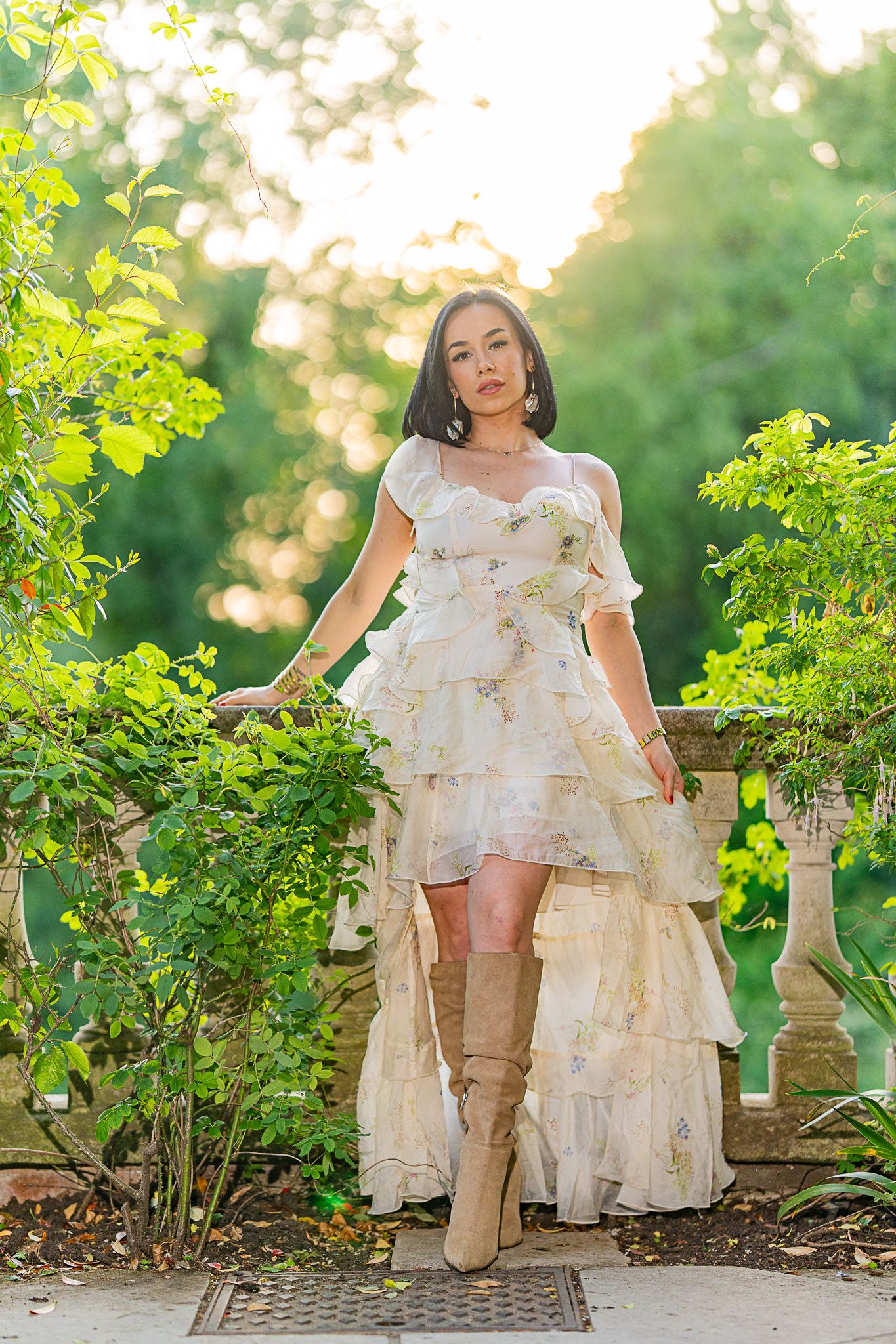 A romantic high-low ruffle dress with tall boots stands elegantly in a sunlit garden setting with stone pillars and greenery.