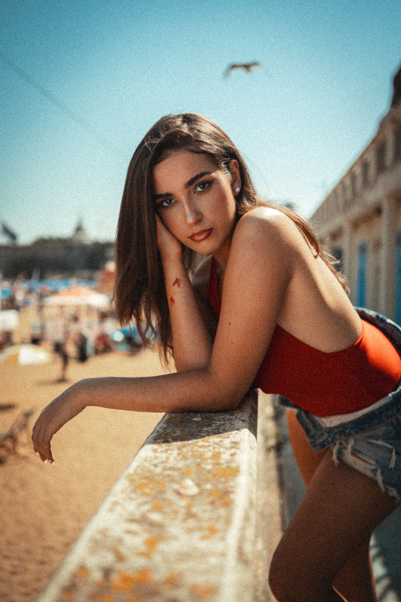 Woman in red swimsuit poses on sunny beach with warm lighting and coastal buildings in background.