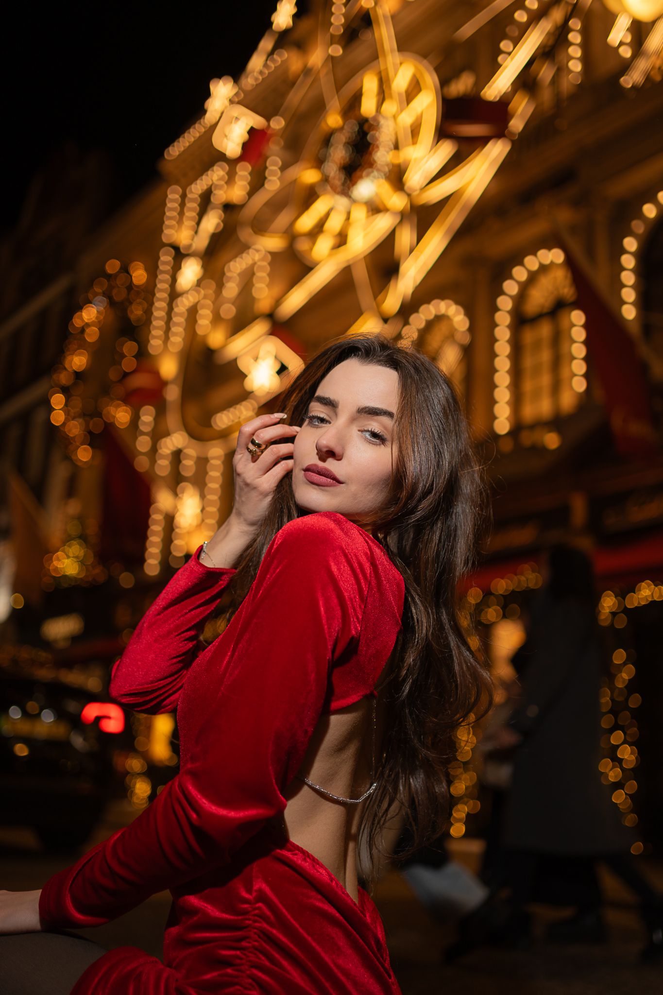 A person in a red velvet dress poses against a backdrop of glowing golden carnival lights at night.