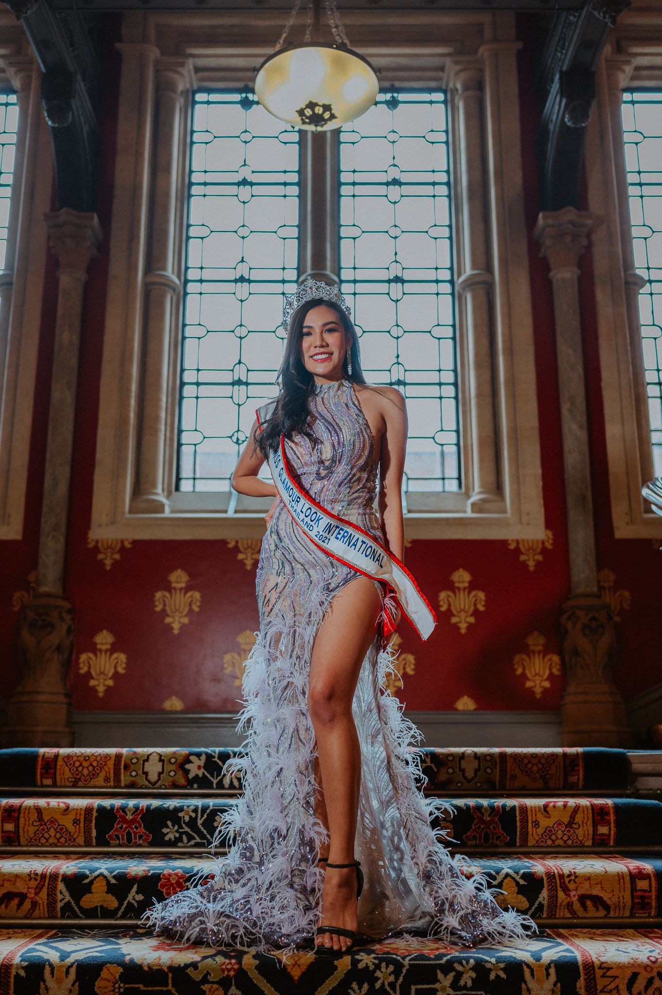 A pageant contestant poses in an elegant silver sequined gown with a high slit in front of a stained glass window.