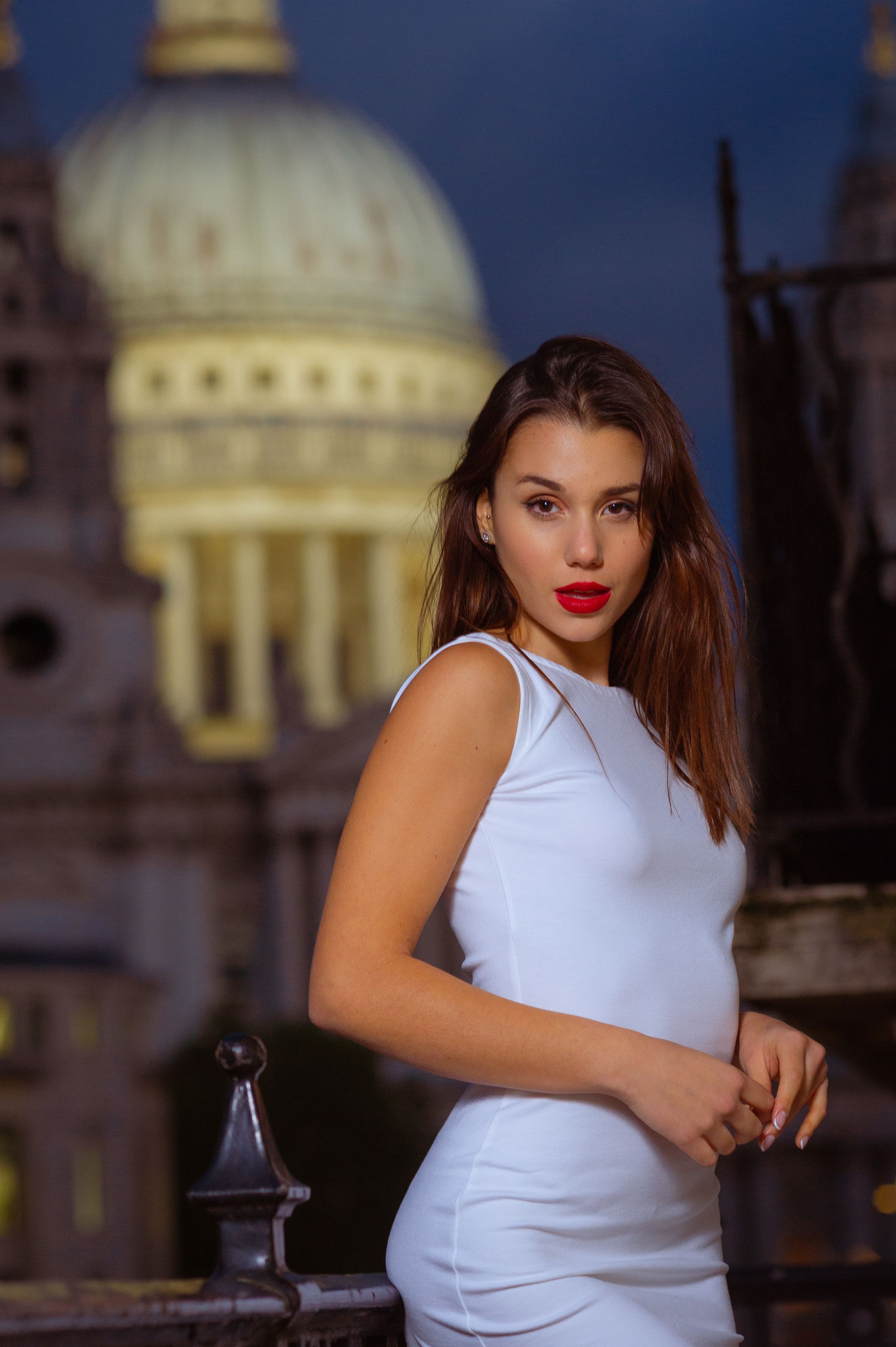 A woman in a white sleeveless dress poses against a dramatic evening cityscape with an illuminated domed building behind her.