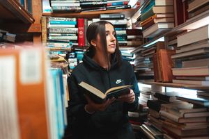 Person browsing through stacks of books in a crowded library with tall wooden bookshelves filled to capacity.