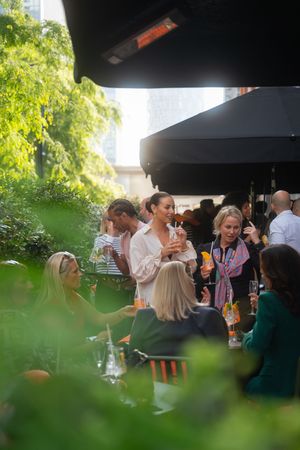People gathered at an outdoor event under a canopy, surrounded by green foliage and natural lighting.