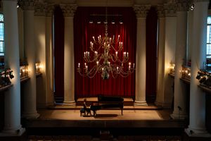 Grand piano sits on stage in ornate concert hall with red curtain backdrop and crystal chandelier hanging above.