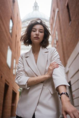 A stylish woman in a beige blazer poses in an urban alley with brick walls and a domed building in background.
