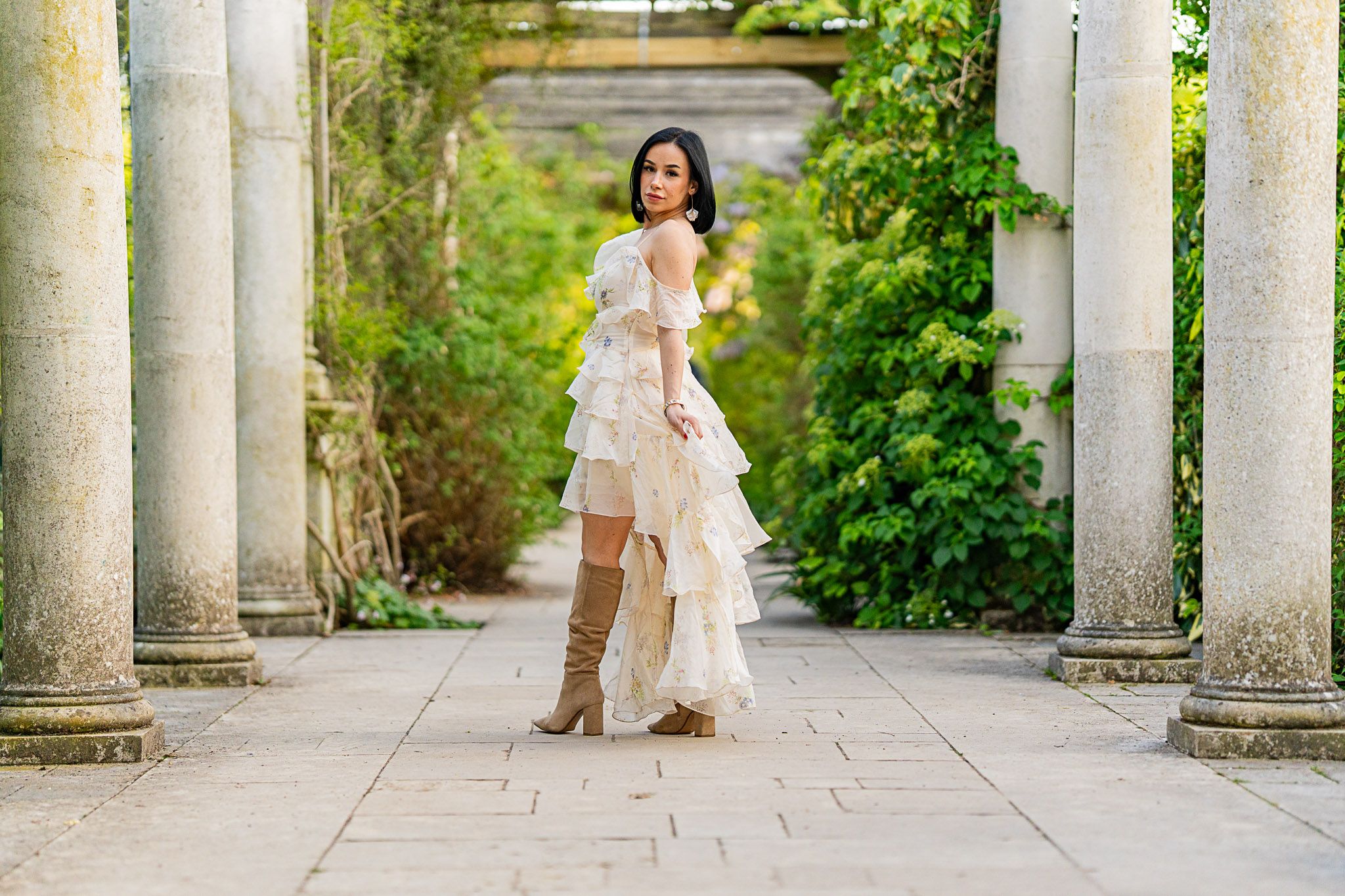 A couple poses for a wedding photo beneath elegant white stone columns in a lush garden setting.