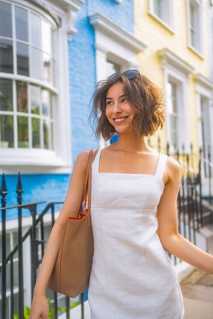 A woman in a white summer dress smiles while walking down a colorful street with pastel buildings and carrying a brown bag.