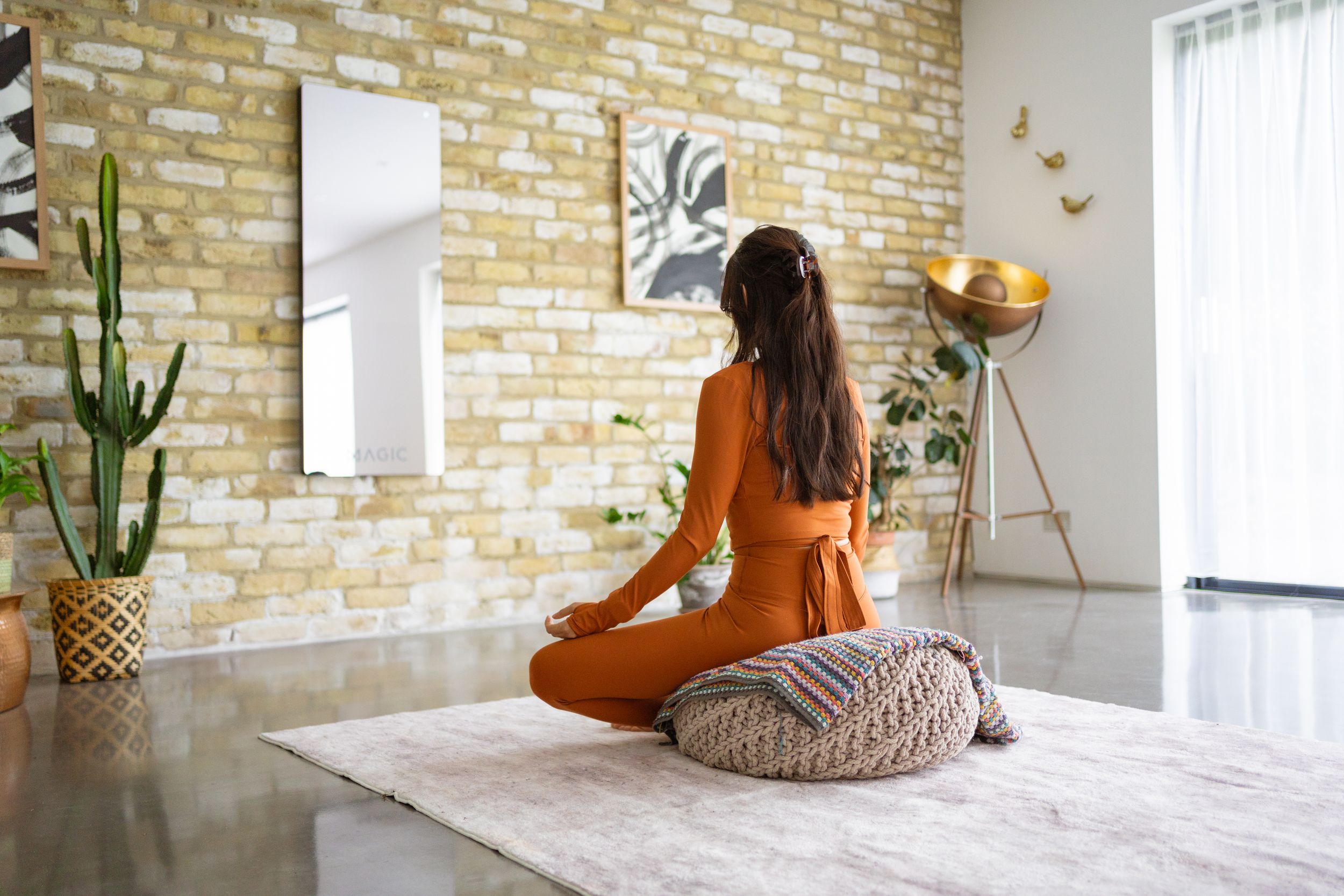 Someone in orange clothing sits on a round cushion near a wall with exposed brick and indoor plants.