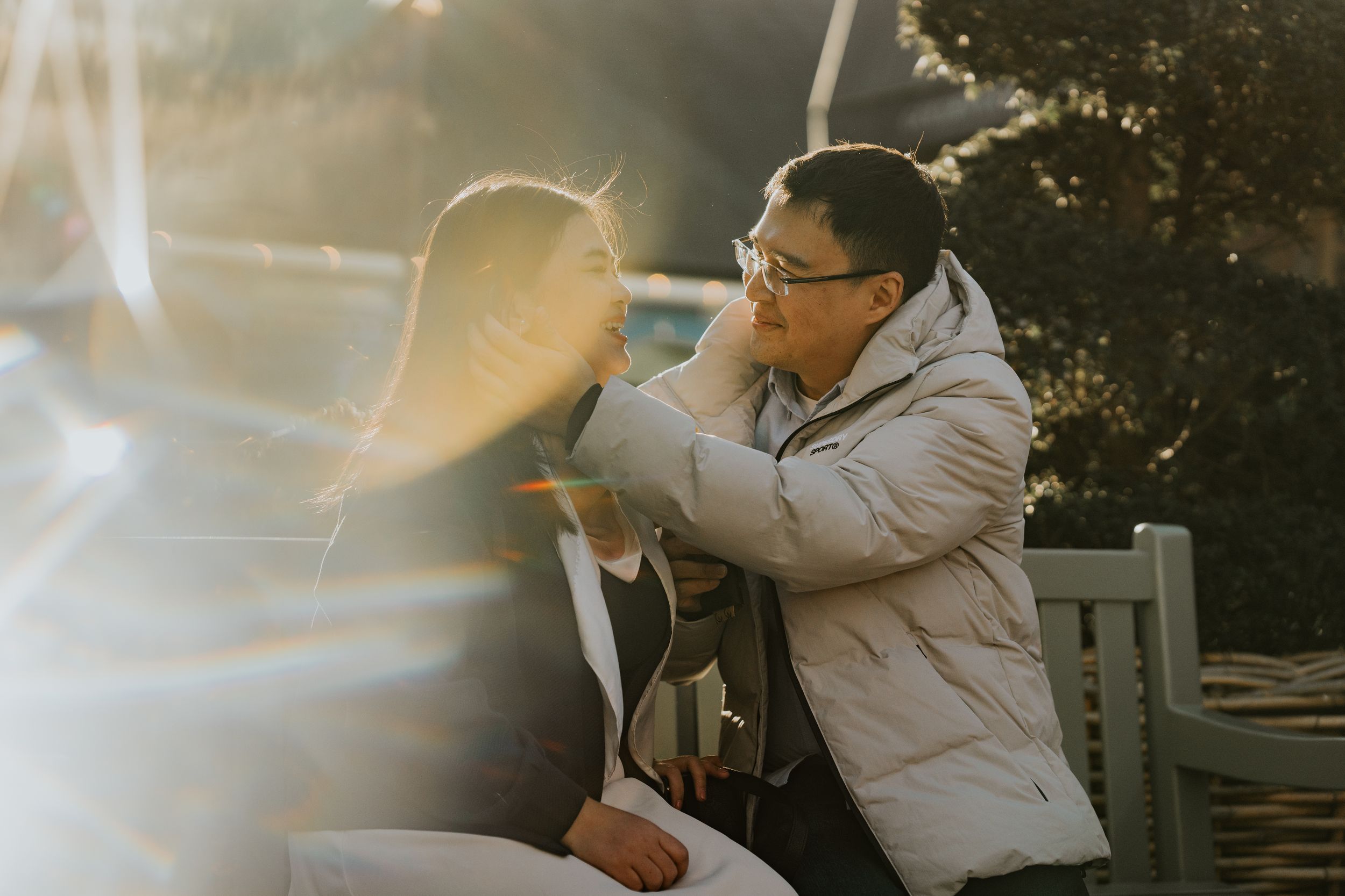 A person wearing a puffy beige jacket sits on a bench during golden hour with sunlight creating lens flare effects.