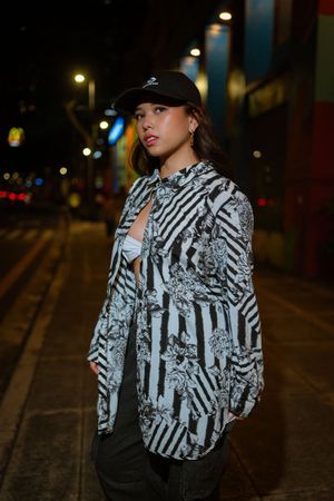A person wearing a black and white striped patterned jacket poses on a dimly lit city street at night.
