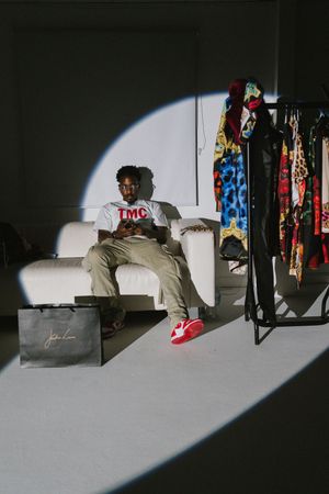 A person sits on a white couch in a spotlight beam beside a clothing rack and shopping bag in a dark room.